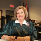 Judge Davenport ousted: Middle-aged white woman with blonde bobbed hair wearing white blouse with black judges' robe sits at podium with microphone in front of dark wood bookcases