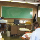 Prison detainees work at desk in a prison classroom with a chalkboard.