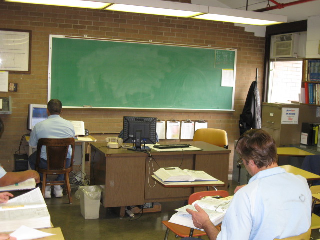 Prison detainees work at desk in a prison classroom with a chalkboard.