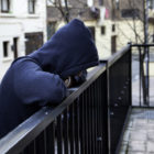 Person in a dark, hooded sweatshirt leans on a black outdoor fence railing,