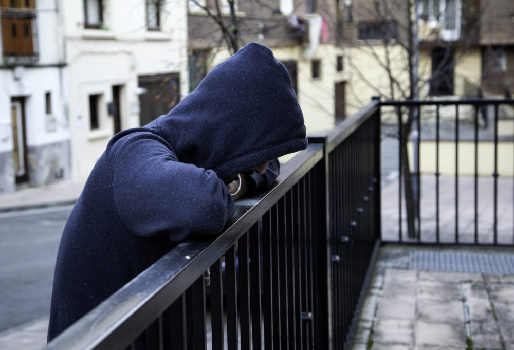 Person in a dark, hooded sweatshirt leans on a black outdoor fence railing,