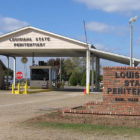 Angola prison: The entrance to the Louisiana State Penitentiary has a guard house that controls entry into the compound with seop sign, three white buildings and brick sign —the sign says "Louisiana State Penitentiary" and "Burl Cain, Warden"