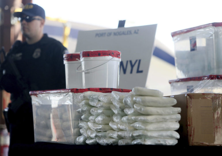 Youth drug overdoses: Clear plastic bags of white powder are stacked on a table with man in dark uniform and baseball cap standing in background