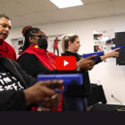 Permitless Carry Georgia: Inside a white-walled classroom, t hree women hold blue training guns at shoulder height aiming in front of themselves while male instructor stands behind them.