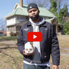 Gun violence survivor campaign: Black man in jeans, a gray winter jacket and navy baseball cap on backwards stands on sidewalk in front of s 2-story oldr home holding a white human skull with large section of bone missing on right side.