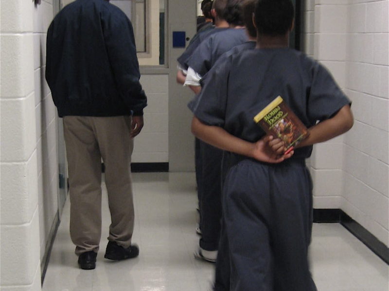 Youth in a blue prisoner jumper holds a book behind his back and is the last in a line of juveniles who are at a detention center get books as a part of a summer reading program.