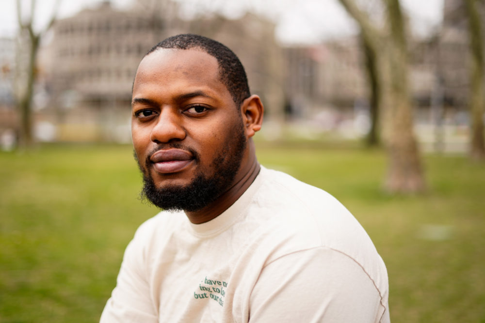 Juvenile Justicee Reform: Black man with short black hair and beard wearing white t-shirt with green grass and trees in the background