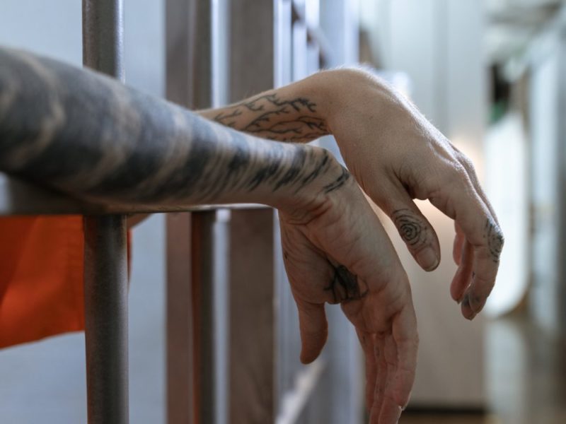 Tattoed forearms and hands rest on cell bars.