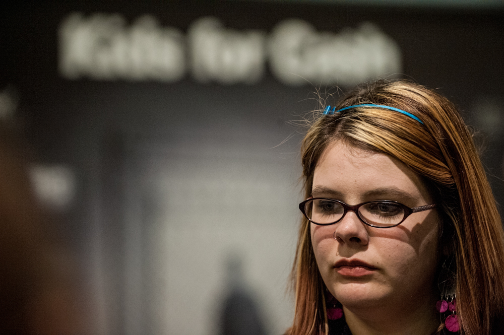 Kids-for-Cash: Headshot of older female teen with blonde hair wearing dark-framed glasses in front of blurred sign saying "Kids for Cash".