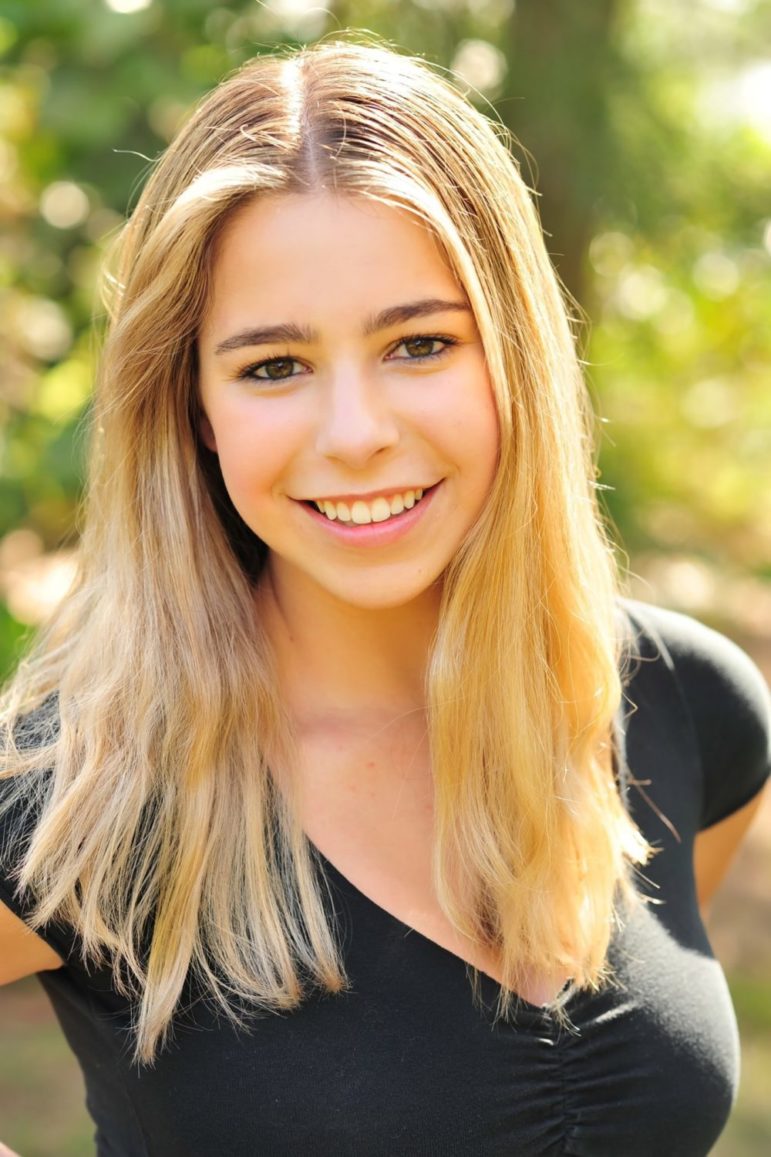 School triage training: Headshot ling-haired blonde teen girl in black top smiling into camera