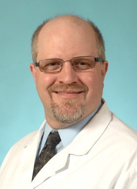 School triage training: Headshot of balding man with glasses in white doctor's coat, blur chirt & dark tie smiles into camera