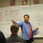 Utah College Classes: Man in blue shirt gestures speaking to clas stanng in front of a white board filled with text