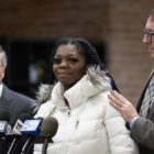 Lawsuit; Illinois agency wrongfully imprisoned children: young black woman in white coat talking into microphones with men in suits at her side