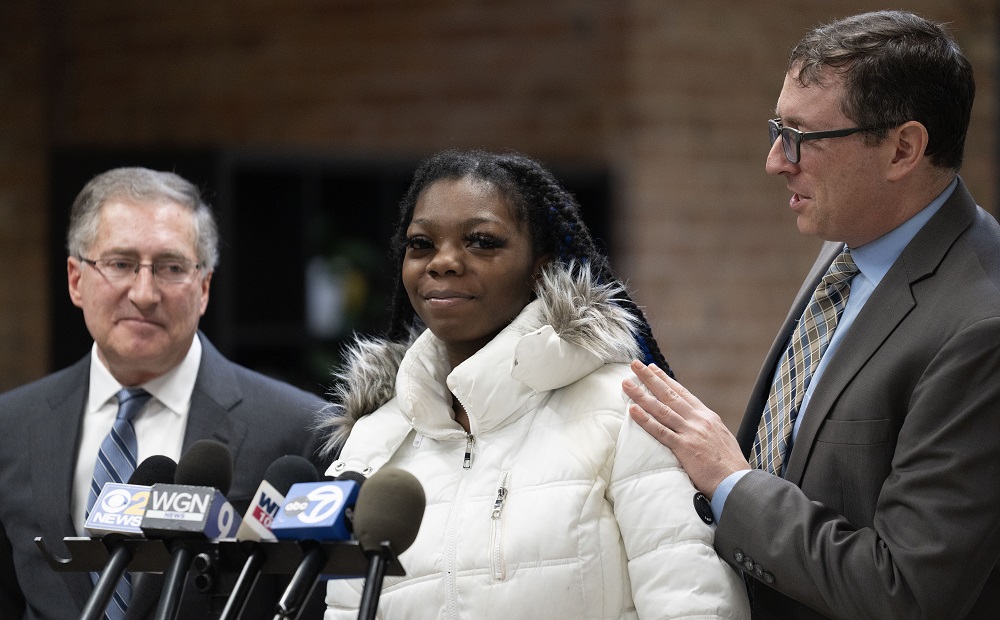 Lawsuit; Illinois agency wrongfully imprisoned children: young black woman in white coat talking into microphones with men in suits at her side