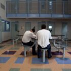 California: Young men sit srpind table with built-in chairs in large ron and stairs leading ro balcony above with doors to several rooms.