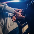 Drug court: Closeup under night streetlighting of policeperson in dark uniform handcuffing a suspect in gray sweatshirt