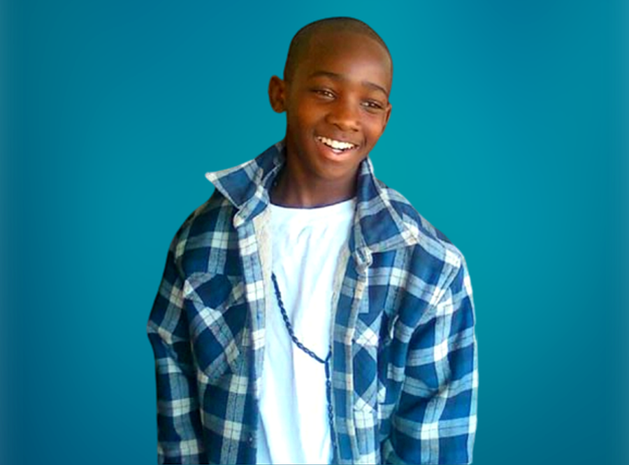 LaKeith Re-Sentencing: Young, black male teen with very short black hair in blue and white plaid long-sleeved shirt over white t-shirt stands in front of turquoise wall smiling broadly into the camera.
