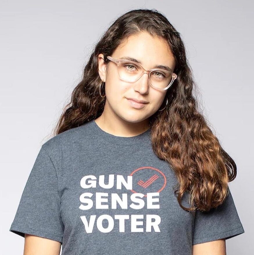 Gun Violence: Older teen with long brown hair and light-framed glasses in gray t-shirt with words "Gun Sense Voter" stands looking into camera