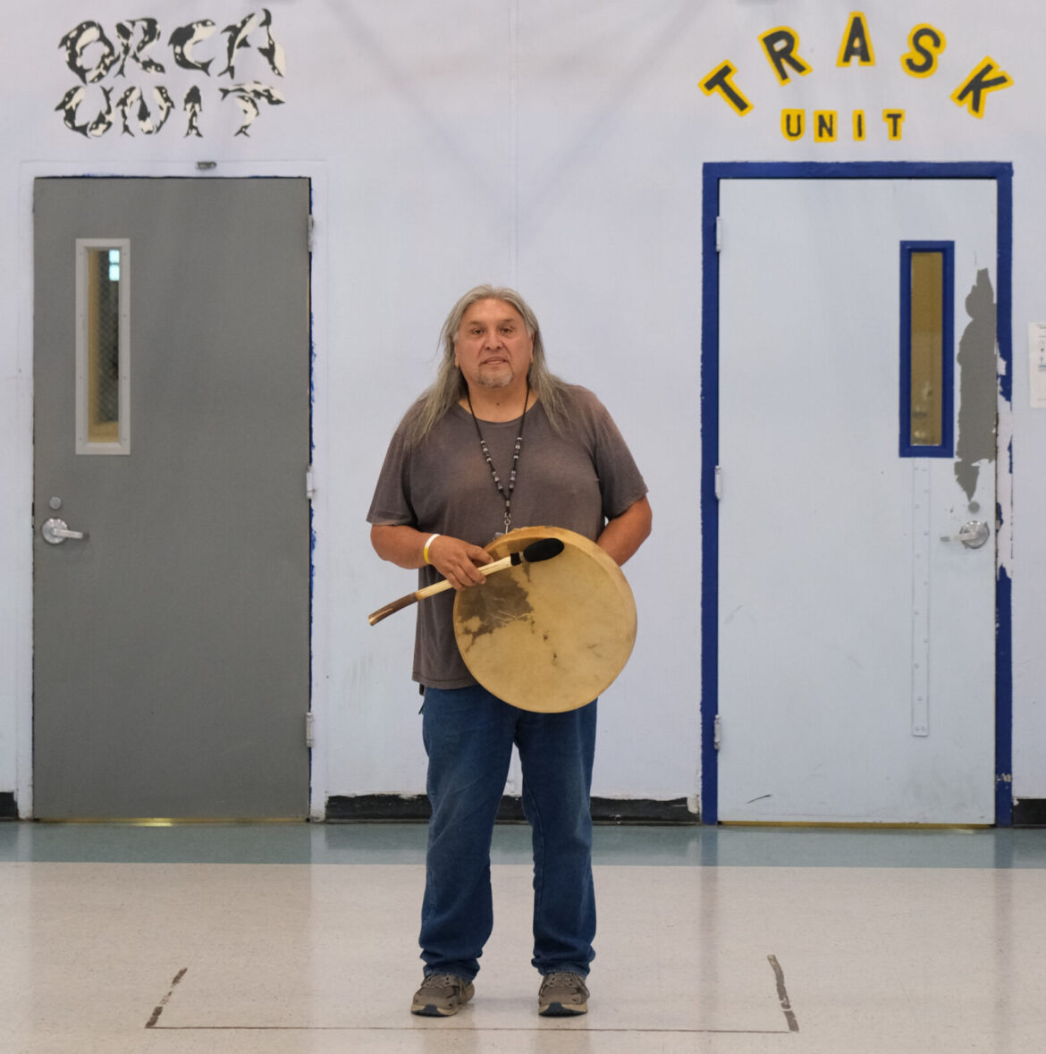 Native American rituals: Native American elder man with long gray hair in jeans and gray t-shirt stands under indoor basketball court hoop holding drum
