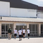 Texas juvenile detention: One adult and four youths wearing black shorts and white t-shirts and ne adult walk toward doors of modern building