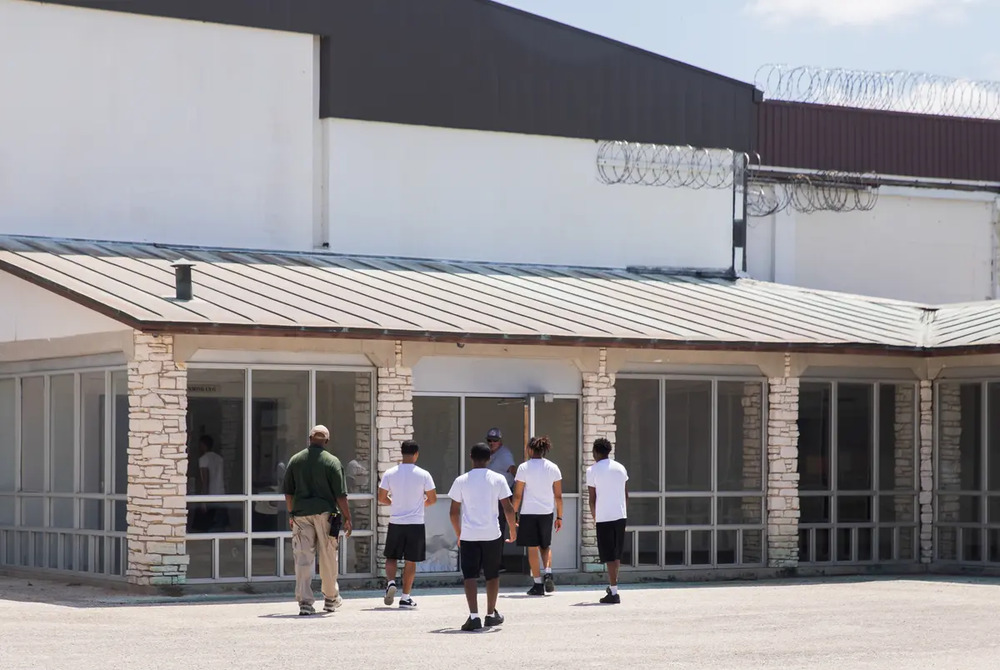 Texas juvenile detention: One adult and four youths wearing black shorts and white t-shirts and ne adult walk toward doors of modern building