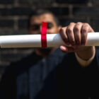 Prison college: Man burred in background wears college graduation gown holding rolled up white paper tied with red ribbon towards camera