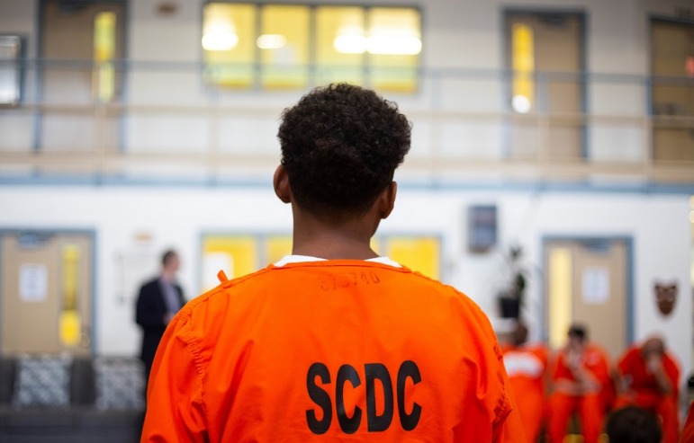 Youth In Adult Prisons: young black prisoner with back to camera in orange prison uniform looking at white wall of doors and interior windows