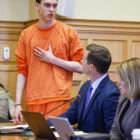Teen murder sentencing: Willard Miller in orange top and pants stands with hand on chest next to adult man and woman seated at a desk who are looking up at him