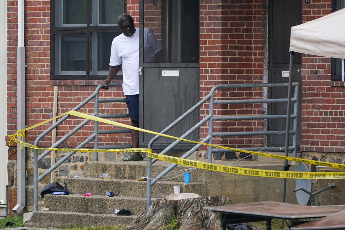 Mass shooting Baltimore: Black adult in white shirt & dark pants stands holding door open on cement porch with stairs in a red brick building