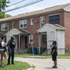 Mass shooting Baltimore: Black policeman in dark uniform stands with 2 black adults outside on grass in front of two-story, red brick apartment building with white porch shelters & trim.