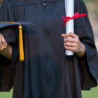 Formerly Incarerated in College: Closeup of person in black graduation gown holding a black graduate cap with gold tassel and a rolled parchment scroll with red ribbon