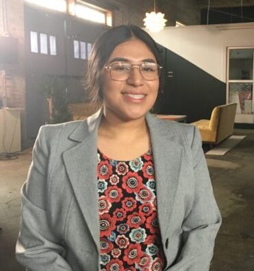 Juvenile justice reform: Young adult Latina woman with black hair pulled back and rimless glasses in light gray suit jacket with red print top stands in warehouse smiling into camera.