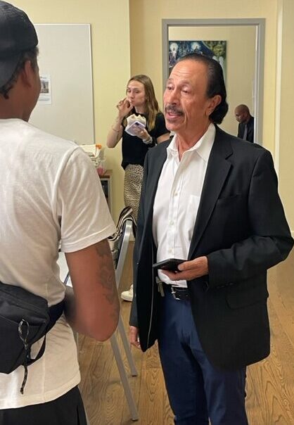First-time juvenile offenders: Older man with dark greying hair and mustache in dark suit and white shirt stands talking to person with back to camera wearing white t-shirt and dark cap.