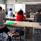 Classroom disruptions: Rear view of several students at desks bent over work with teacher facing classroom standing in front of blackboard.