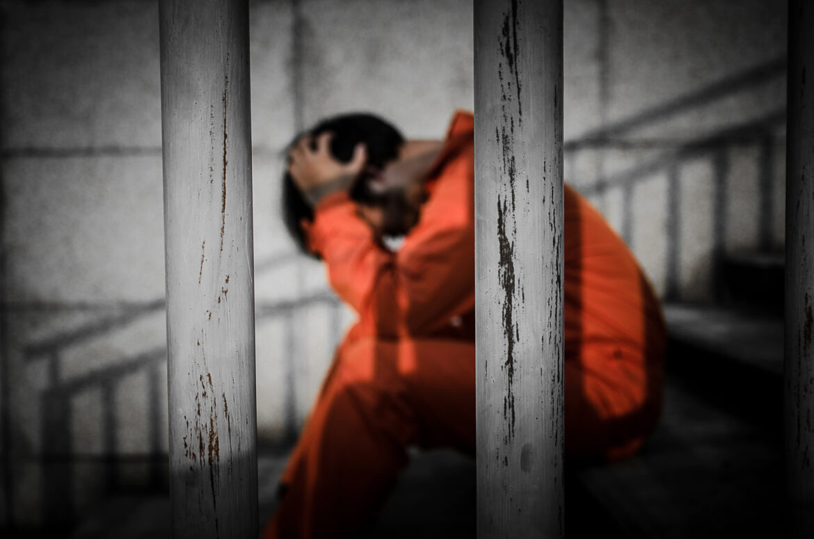 Juvenile justice reform Louisiana: Person with dark hair sits on steps behind bars with head in hands wearing orange jumpsuit