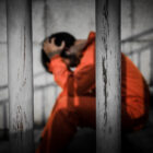 Juvenile justice reform Louisiana: Person with dark hair sits on steps behind bars with head in hands wearing orange jumpsuit