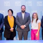 Mentoring month: 3 men and 2 women in business cloths stand in front of floor to ceiling light blue screen with white Hope Florida logo repeat printed across several rows