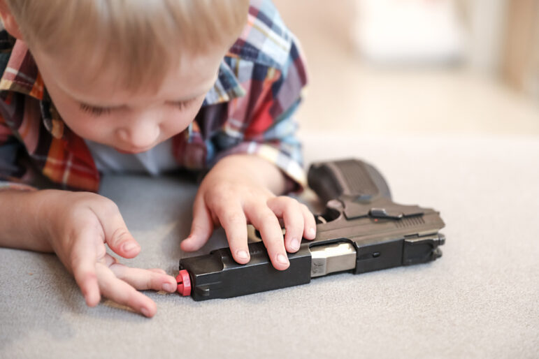 Gun Safety ar Home: Very young boy leans close to hand fun on table poking one finger into barrel.
