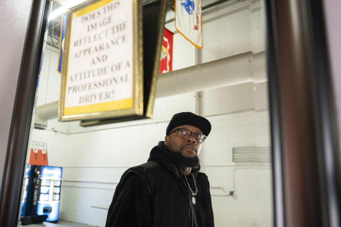 Prison No Education: Black man in dark winter jacket, hat stands inside large, white-walled warehouse space with very high ceilings. Above his head are signs hanging from ceiling and soda vending machines in background.