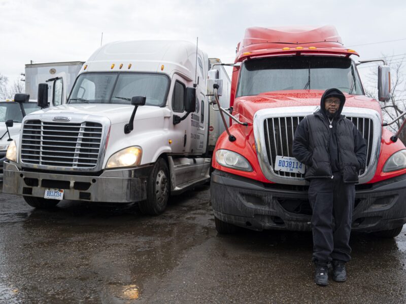 Prison No Education: Black man in dark winter jacket, hat and pants stands leaning against front of bright red semi-truck parked next to a white semi-truck on asphalt parking lot under gray, rainy sky