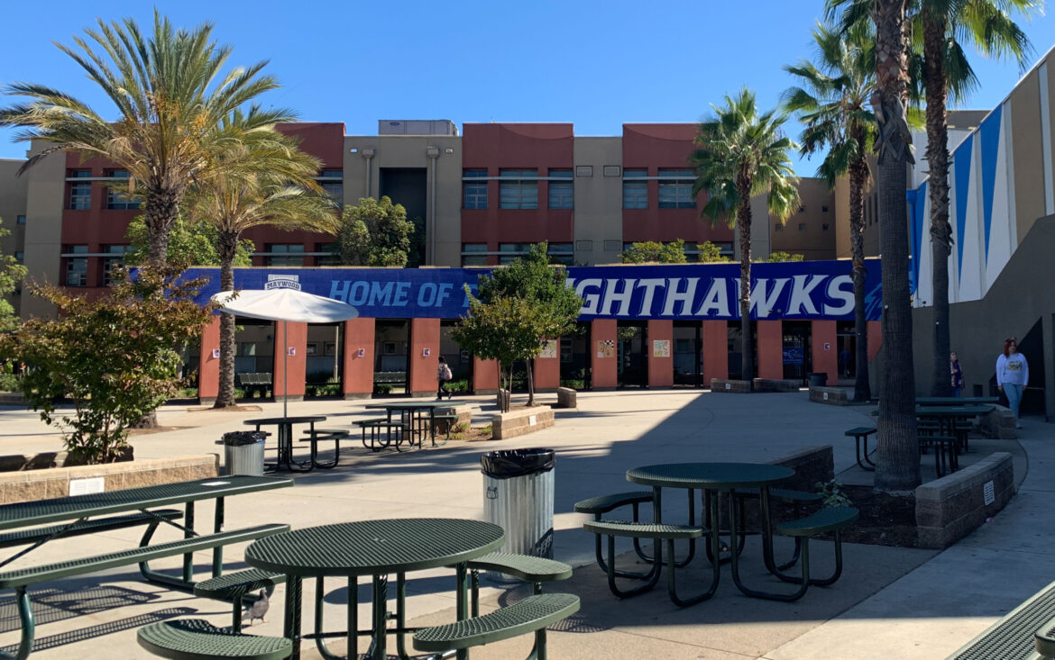 No school suspension: Several empty round tables with attached benches in empty cement courtyard with three-storty red brick building in background hung with white text-on-blue banner saying "Nighthawks"