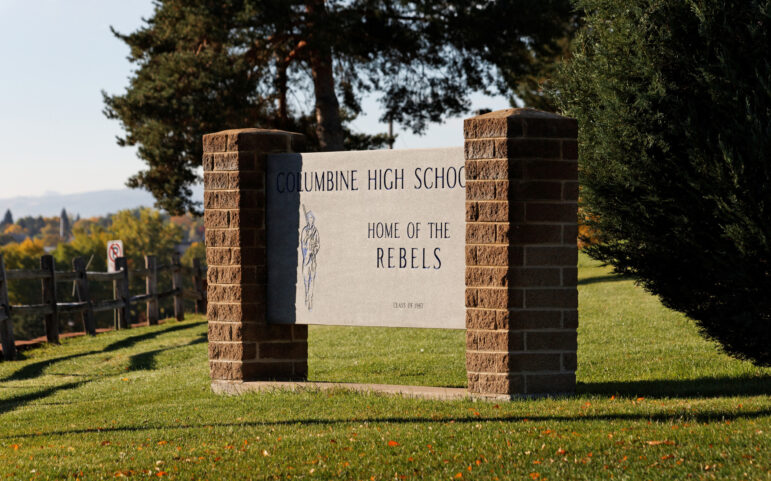 Q&A Teaching after Columbine: Columbine High School sign - cement center with red brick pillars - in 2012, on grass slope with evergreen trees in background.