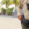 Police on campus: closeup of only middle and right side body showing hand on gun in holster of police on tan and brown uniform with modern high school campus in the background