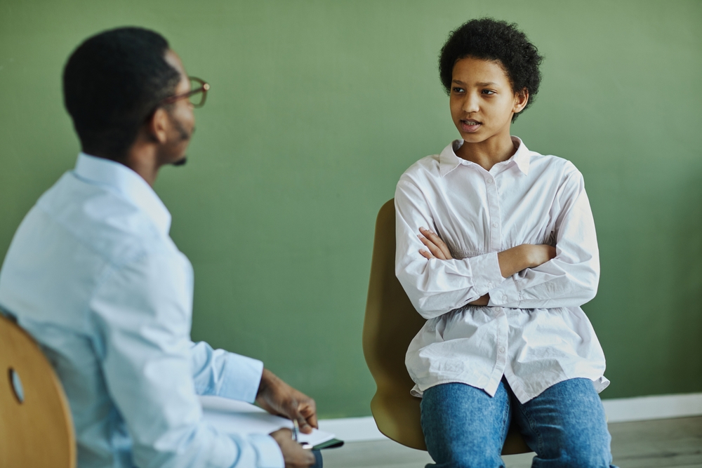 Schools and teen violence: Black male teen sits sullenly in classroom chair listening to Black male teacher both in front of green chalkboard