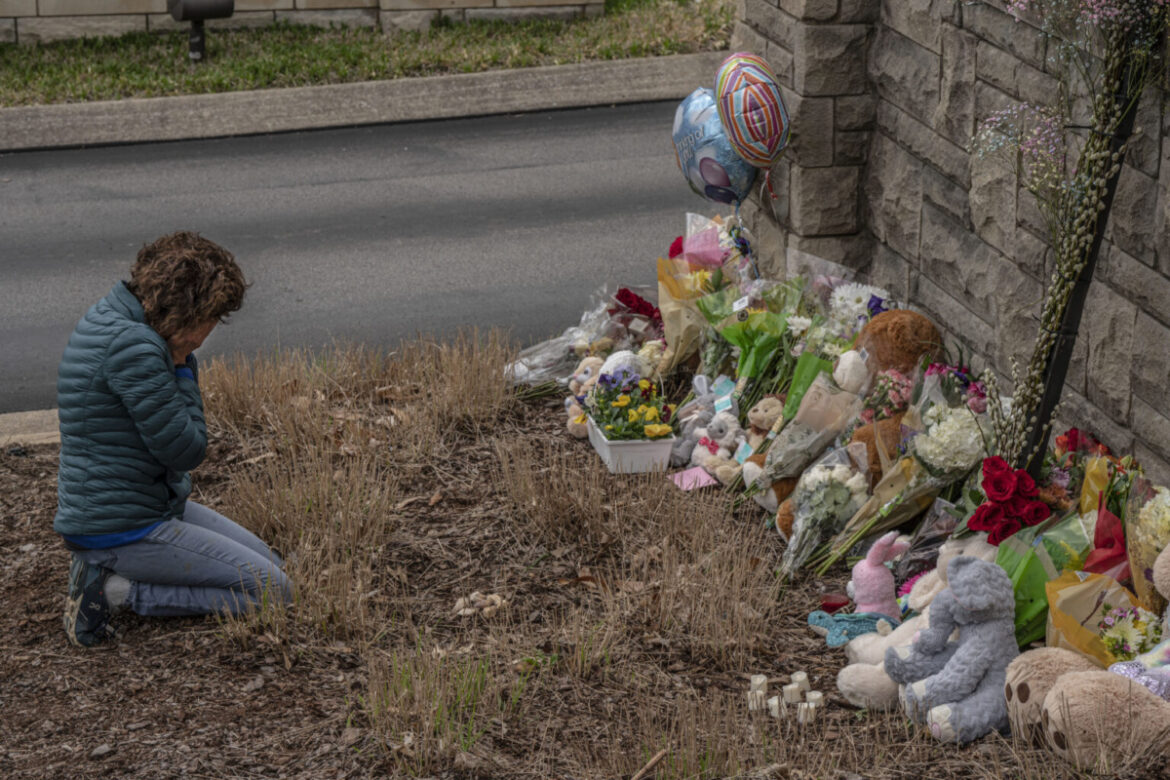 Tennessee Schools: Person with short brown hair with head down and hands covering face kneels in dry grass in front of stone wall with flowers, stuffed animals and balloons piled up against it