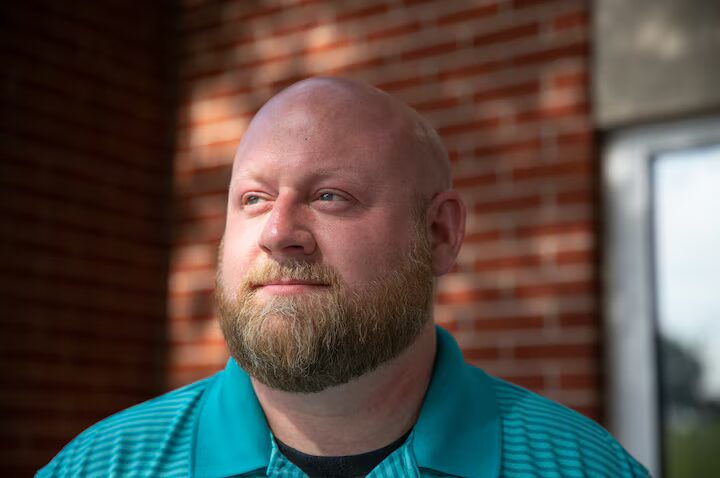 Arming teachers in Tennessee: Headshot of bald white man with light brown full beard in teal and blue striped polo shirt in front of red brick wall