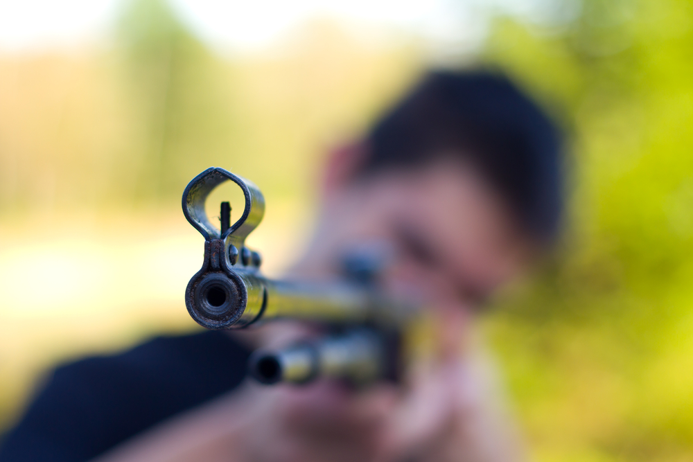 Cruz no death penalty: Blurred teen male head with blurred trees background holding and aiming a long gun pointed at the camera