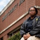 Teens as Texas Jail Guards: Teen girl in tan pants and dark uniform jacket sitting outside a red brick building on a low red brick entryway wall