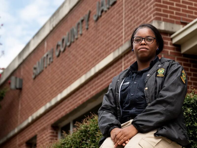 Teens as Texas Jail Guards: Teen girl in tan pants and dark uniform jacket sitting outside a red brick building on a low red brick entryway wall