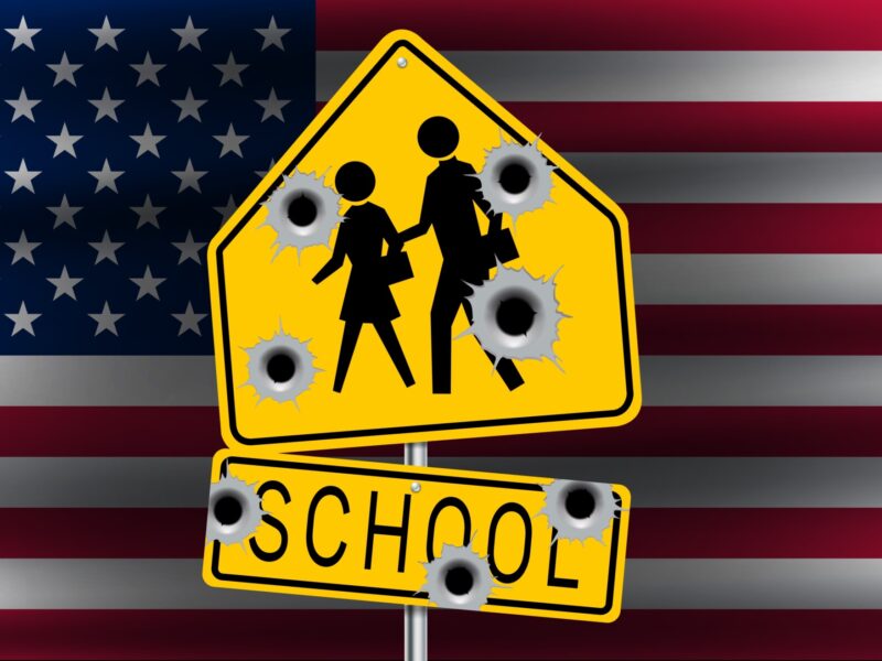 Mass shootings: American flag in background with centered yellow and black school crossing sign with 7 bullet holes.